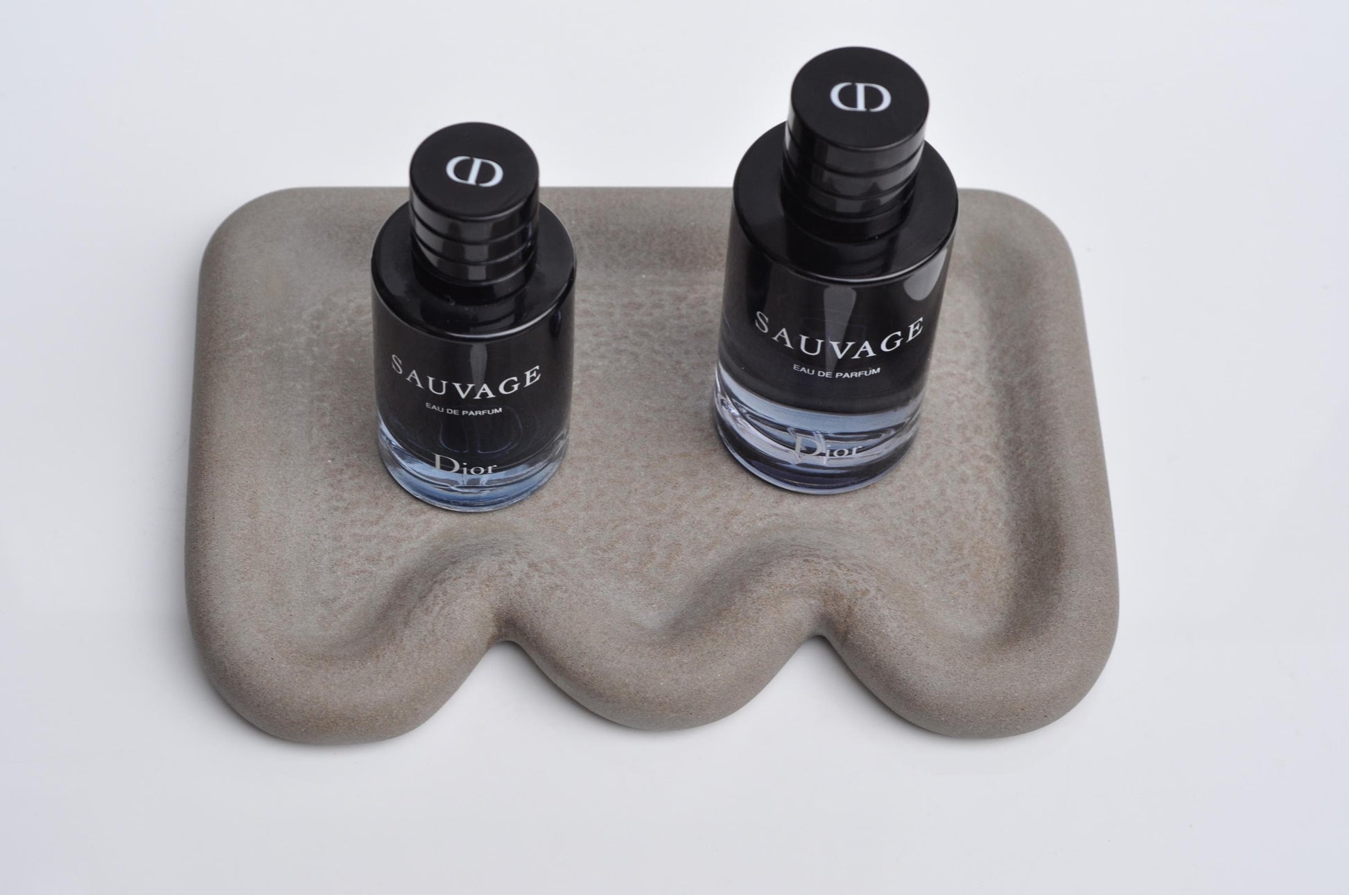 Two bottles of Dior Sauvage cologne on a wavy stone surface with a white background