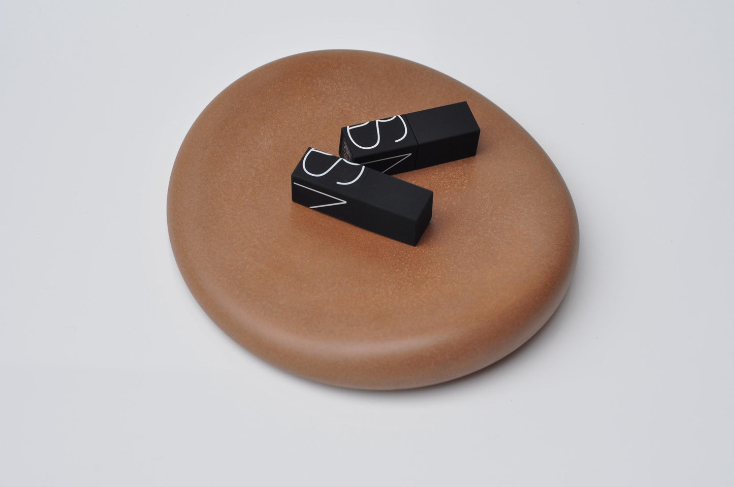 Two black makeup products on a brown circular tray against a white background