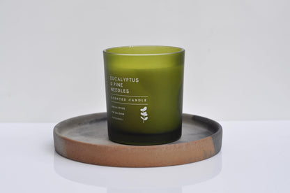 Green candle labeled 'Eucalyptus & Pine Needles' on a concrete coaster with a white background